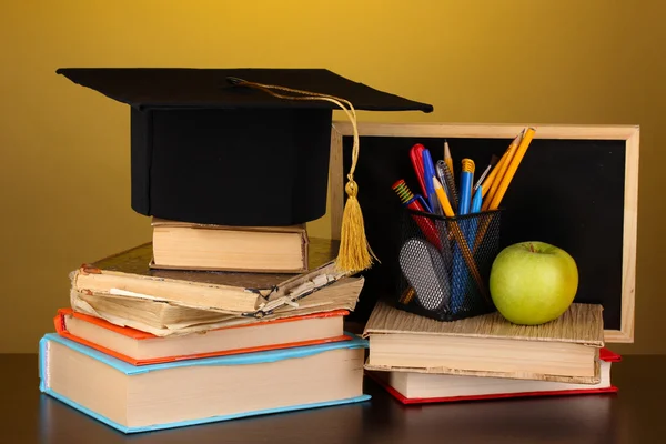 Bücher und Mütze gegen Schultafel auf Holztisch auf gelbem Hintergrund — Stockfoto