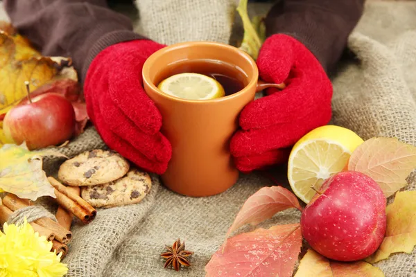 Hands holding cup of hot drink and autumn leaves, on burlap background — Stock Photo, Image