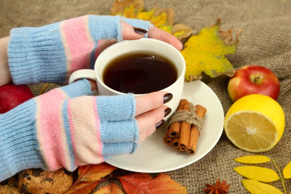 Hands holding cup of hot drink and autumn leaves, on burlap background — Stock Photo, Image