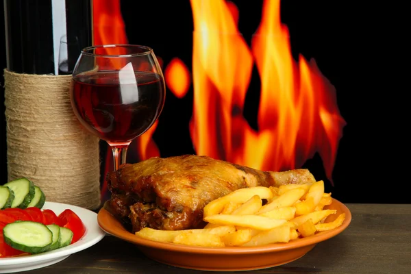 Roast chicken with french fries and cucumbers, glass of wine on wooden table on fire background — Stock Photo, Image