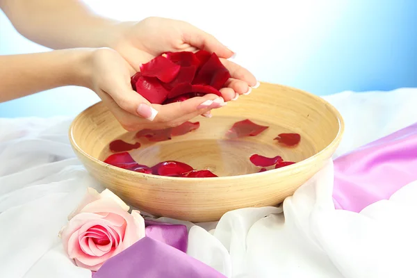 Woman hands with wooden bowl of water with petals, on blue background — Stock Photo, Image