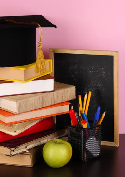 Libros y gorra magister contra pizarra escolar sobre mesa de madera sobre fondo rosa —  Fotos de Stock