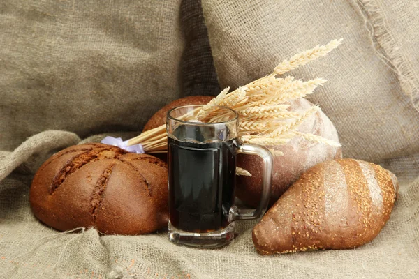 Tankard of kvass and rye breads with ears, on burlap background — Stock Photo, Image