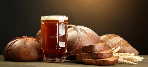 Tankard of kvass and rye breads with ears, on wooden table on brown background — Stock Photo, Image