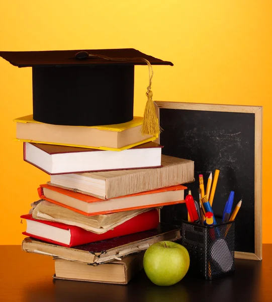 Books and magister cap against school board on wooden table on yellow background — Stock Photo, Image