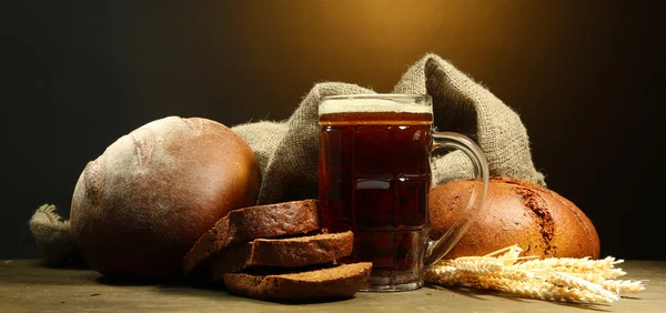 Tankard of kvass and rye breads with ears, on wooden table on brown background — Stock Photo, Image