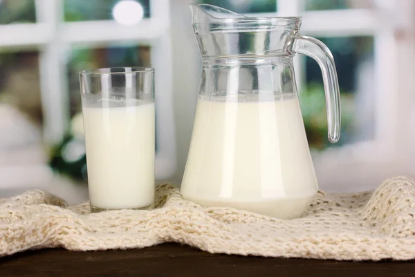 Jarro e copo de leite em tripulações malhas na mesa de madeira no fundo da janela — Fotografia de Stock