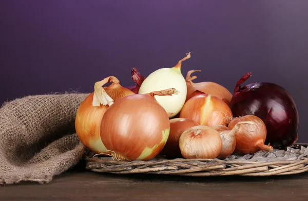 Rijp uien op rieten wieg op houten tafel op paarse achtergrond — Stockfoto