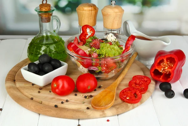 Ensalada griega fresca en un tazón de vidrio rodeado de ingredientes para cocinar en una mesa de madera sobre un fondo de ventana — Foto de Stock