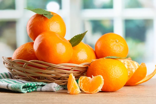 Tangerines with leaves in a beautiful basket, on wooden table on window background — Stock Photo, Image