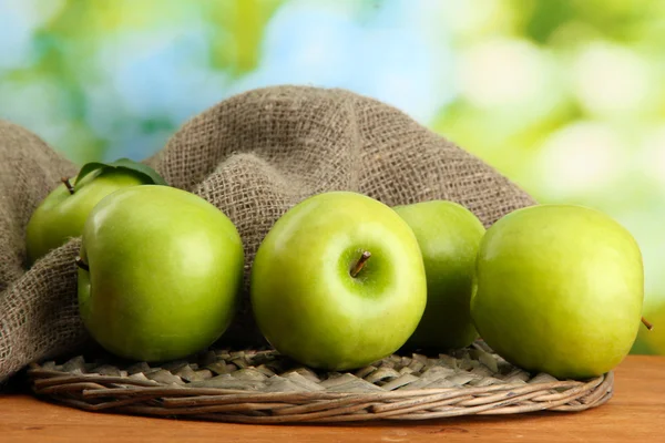 Ripe green apples with leaves on burlap, on wooden table, on green background — Stock Photo, Image