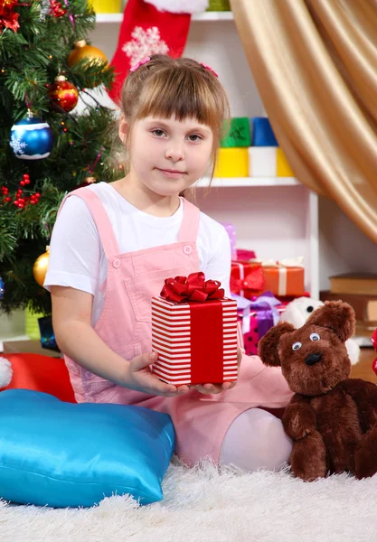Маленькая девочка сидит рядом с елкой с подарком в празднично украшенной комнате — стоковое фото
