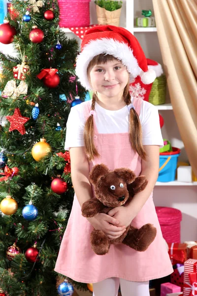 Little girl in Santa hat near the Christmas tree in festively decorated room — Stock Photo, Image