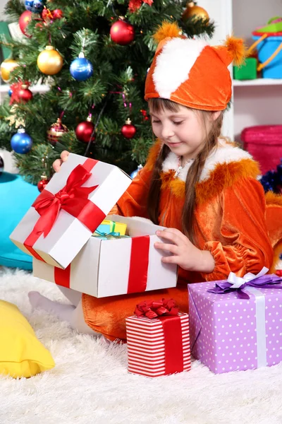 Маленькая девочка в костюме белки открывает подарок в празднично украшенной комнате — стоковое фото