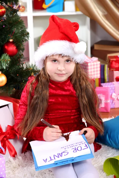 Hermosa niña en vestido rojo escribe carta a Santa Claus en habitación festivamente decorada — Foto de Stock