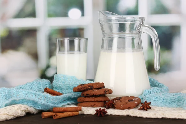 Pitcher and glass of milk with cookies on crewnecks knitwear on wooden table on window background — Stock Photo, Image