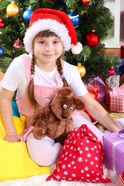 Kleines Mädchen mit Weihnachtsmütze in der Nähe des Weihnachtsbaums in festlich geschmücktem Raum — Stockfoto