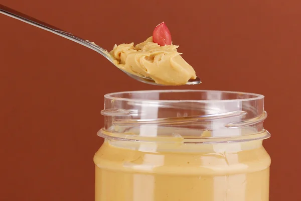 Deliciosa manteiga de amendoim em frasco e colher no fundo marrom — Fotografia de Stock