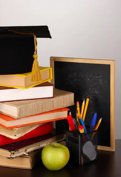 Buku dan topi magister terhadap papan sekolah di atas meja kayu pada latar belakang abu-abu — Stok Foto