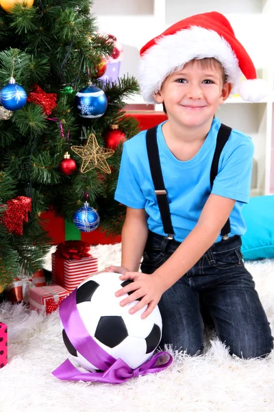 Маленький мальчик в шляпе Санты сидит рядом с ёлкой с футбольным мячом. — стоковое фото