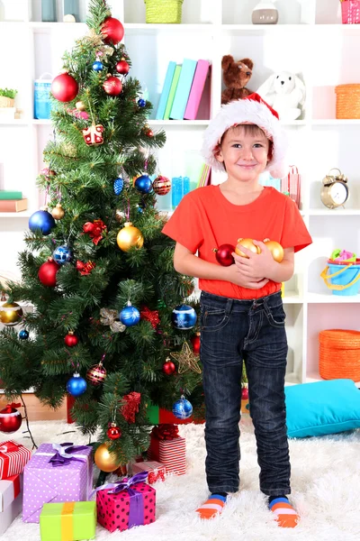 Kleine jongen in Kerstman hoed siert kerstboom op kamer — Stockfoto