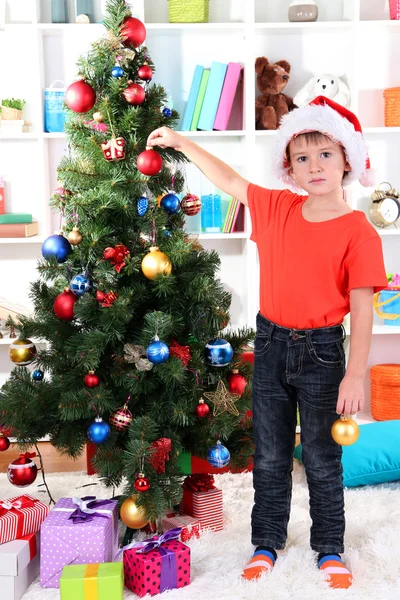 Little boy in Santa hat decorates Christmas tree in room Stock Image