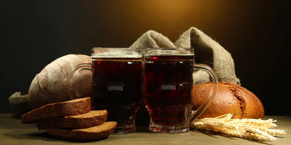 Tankards of kvass and rye breads with ears, on wooden table on brown background — Stock Photo, Image