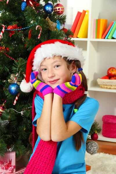Little girl with pink scarf and multicolor gloves sitting near christmas tree — Stock Photo, Image