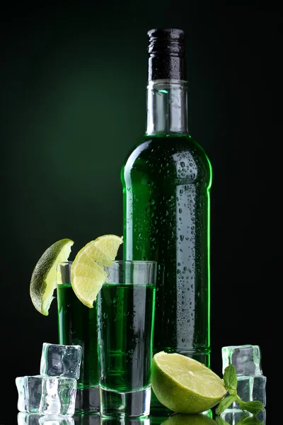 Bottle and glasses of absinthe with lime and ice on green background — Stock Photo, Image