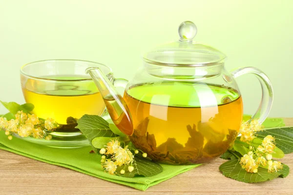 Teapot and cup with linden tea and flowers on wooden table on green background — Stock Photo, Image