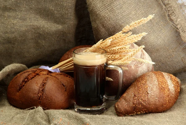 Tankard of kvass and rye breads with ears, on burlap background — Stock Photo, Image