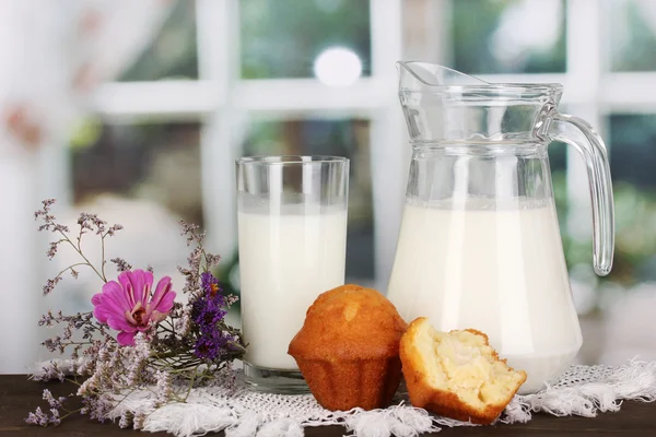 Στάμνα και ποτήρι γάλα με muffins σε ξύλινο τραπέζι σε φόντο παράθυρο — Φωτογραφία Αρχείου