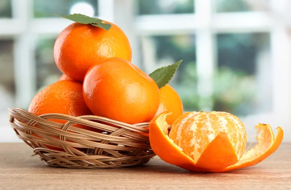 Tangerines with leaves in a beautiful basket, on wooden table on window background — Stock Photo, Image