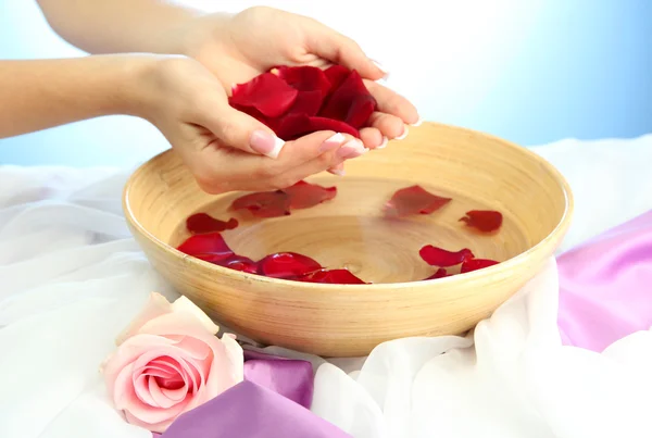 Woman hands with wooden bowl of water with petals, on blue background — Stock Photo, Image