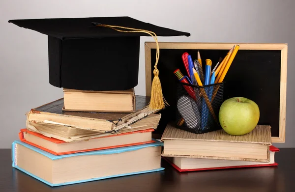 Books and magister cap against school board on wooden table on grey background — Stock Photo, Image