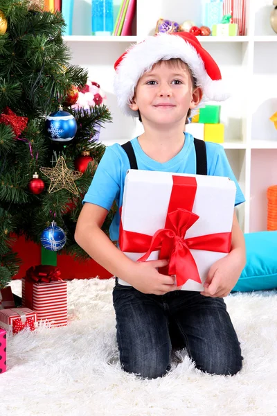 Menino em chapéu de Papai Noel se senta perto da árvore de Natal com presente nas mãos — Fotografia de Stock