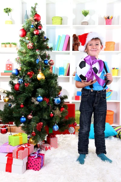 Little boy in Santa hat stands near Christmas tree with football ball Royalty Free Stock Photos