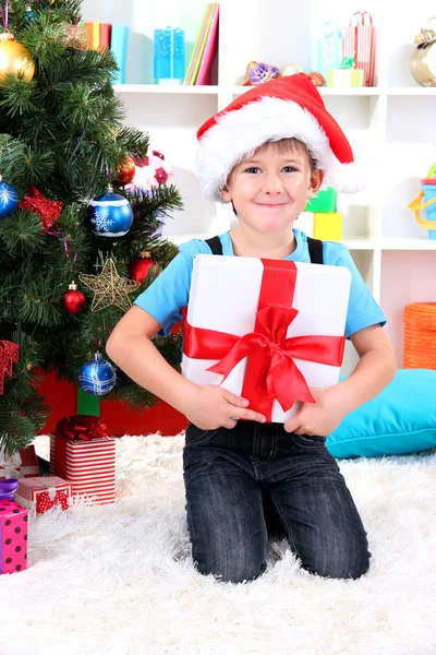 Little boy in Santa hat sits near Christmas tree with gift in hands Stock Photo