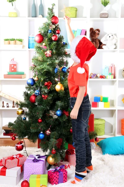 Little boy in Santa hat decorates Christmas tree in room Stock Photo