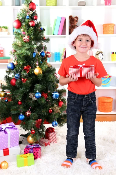 Little boy in Santa hat stands near Christmas tree with gift in his hands Stock Picture