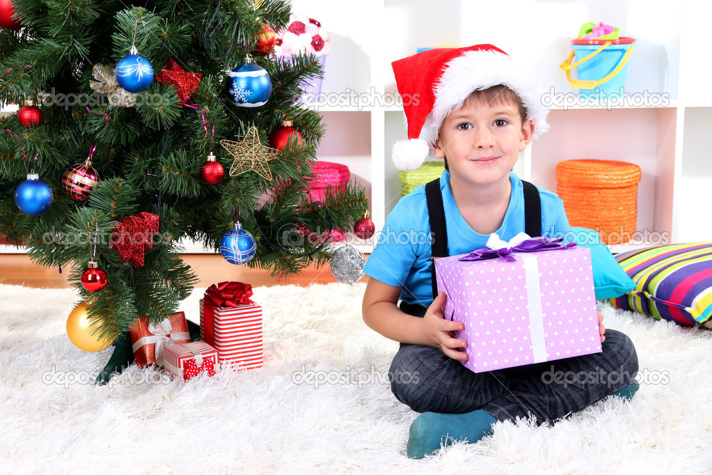 Child in Santa hat sits near Christmas tree with gift in hands
