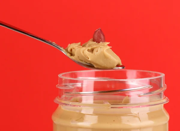 Köstliche Erdnussbutter in Glas und Löffel auf rotem Hintergrund — Stockfoto