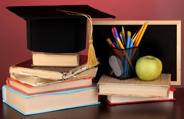 Books and magister cap against school board on wooden table on red background — Stock Photo, Image