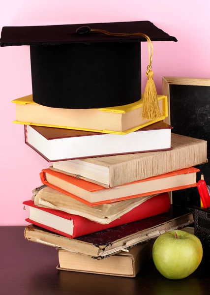 Books and magister cap against school board on wooden table on pink background — Stock Photo, Image