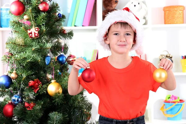 Menino em chapéu de Papai Noel decora árvore de Natal no quarto — Fotografia de Stock