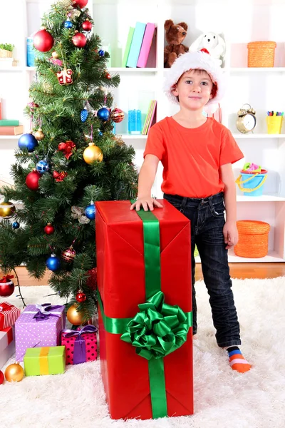 Ребенок в шляпе Санты рядом с елкой с большим подарком — стоковое фото