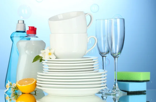 Empty clean plates, glasses and cups with dishwashing liquid, sponges and lemon on blue background — Stock Photo, Image