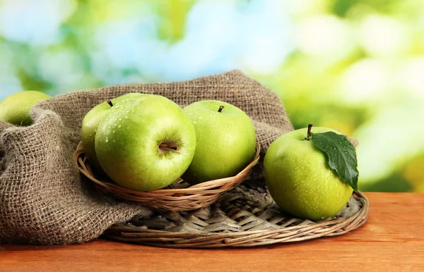 Ripe green apples in basket on burlap, on wooden table, on green background — Stock Photo, Image