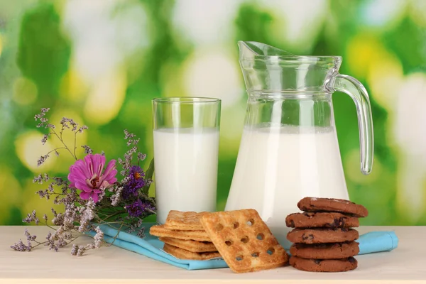 Кувшин и стакан молока с печеньем на деревянном столе на естественном фоне — стоковое фото