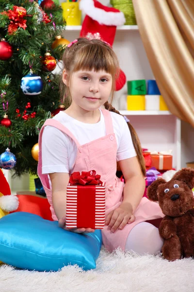 Niña se sienta cerca de un árbol de Navidad con regalo en la mano en la habitación festivamente decorada —  Fotos de Stock